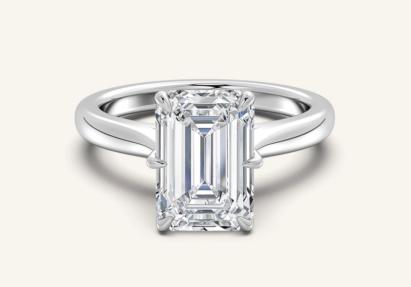 An emerald-cut diamond engagement ring with a polished silver-colored band, shown on a light cream background.